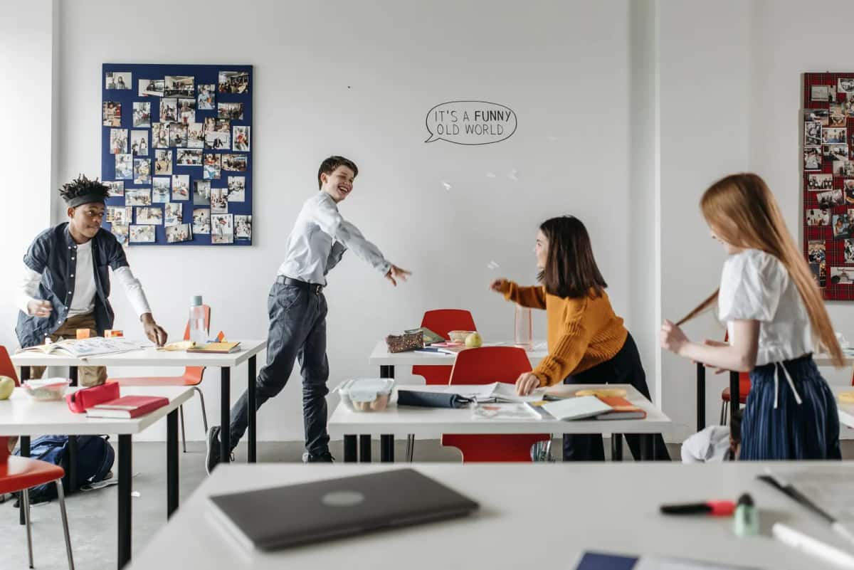 Salle de classe lumineuse où cinq adolescents et leur professeur travaillent debout et assis autour de tables blanches