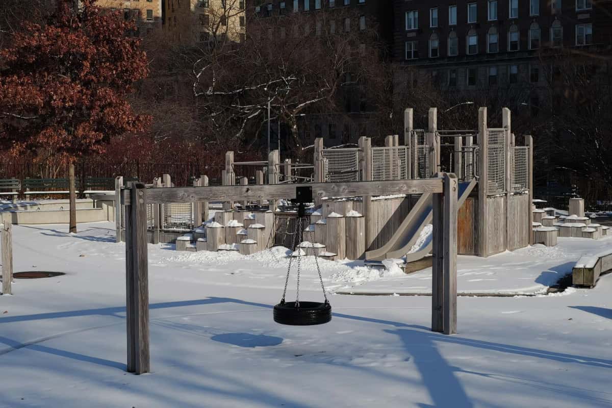 Aire de jeux en bois sous la neige, balançoire pneu et tour de jeux devant les façades d'un quartier résidentiel