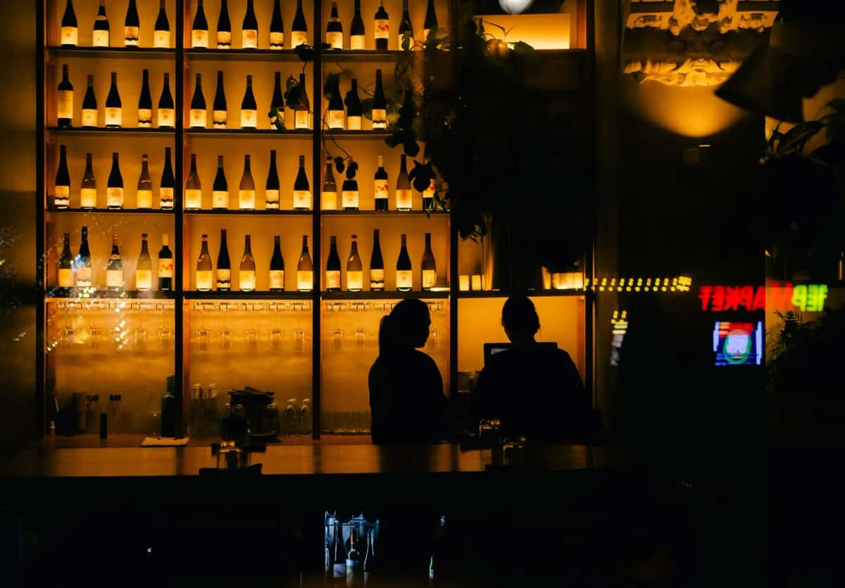 Comptoir de bar le soir, étagères de bouteilles rétroéclairées et deux clients en silhouette devant la vitrine