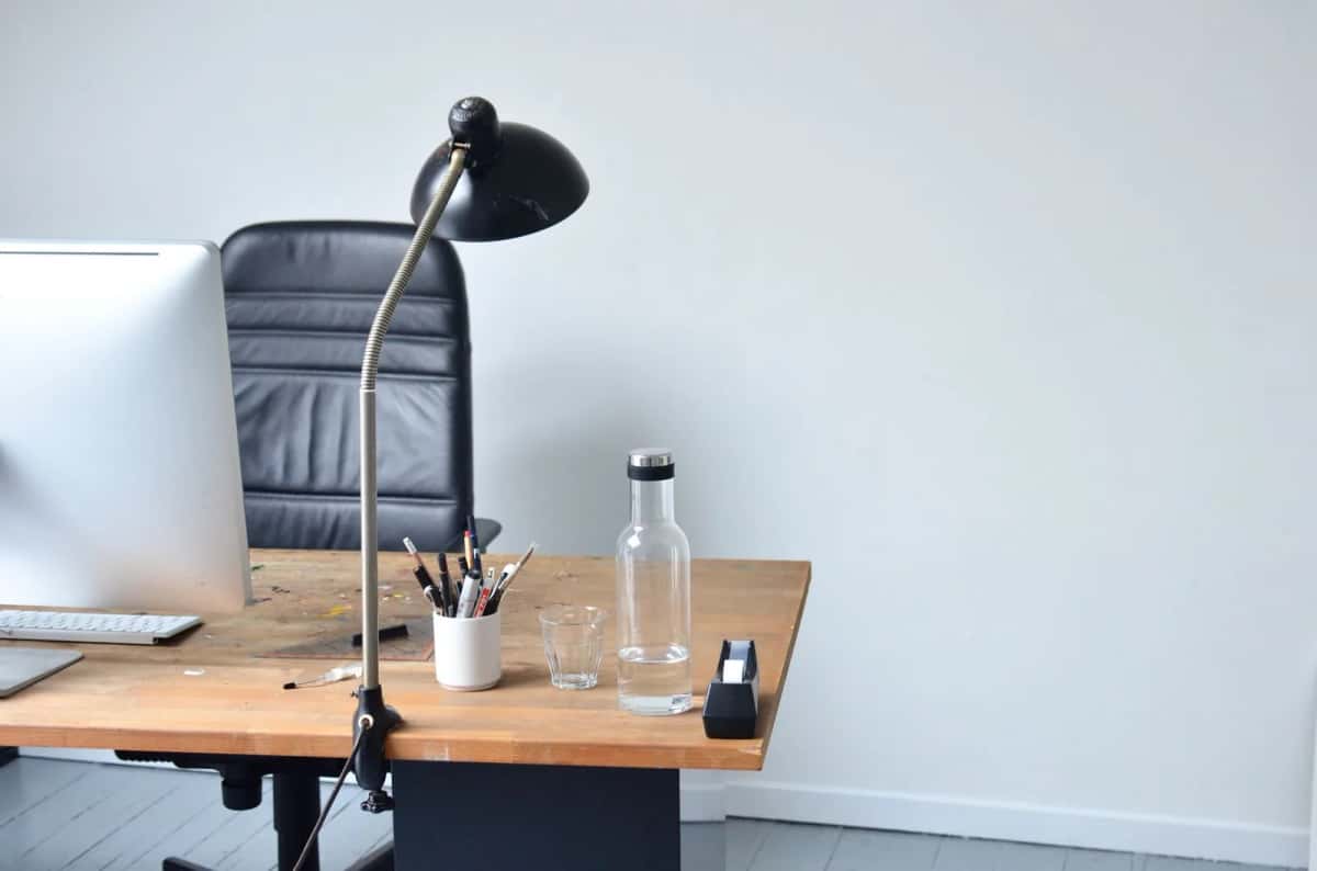 Un bureau clair avec un grand écran, une lampe articulée noire, une carafe d'eau et un fauteuil de travail