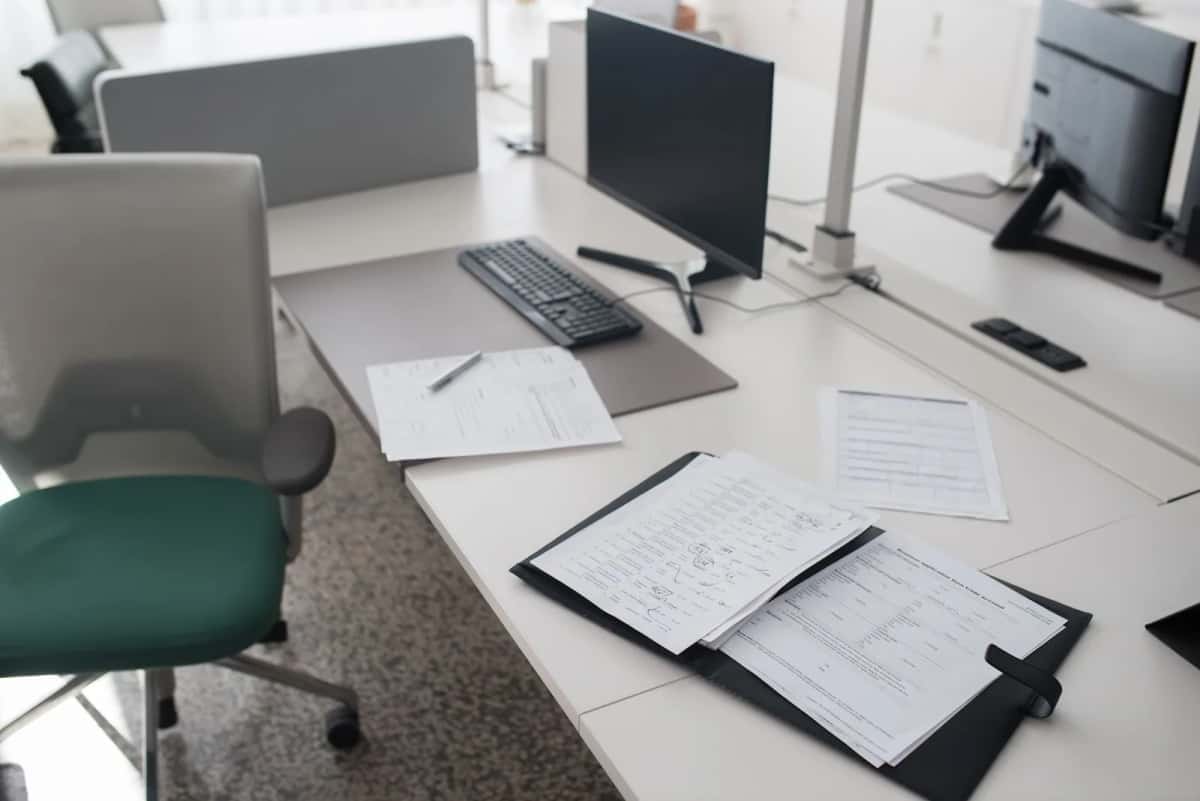 Plusieurs postes de travail vides dans un bureau en open space, écrans éteints et documents posés sur les tables