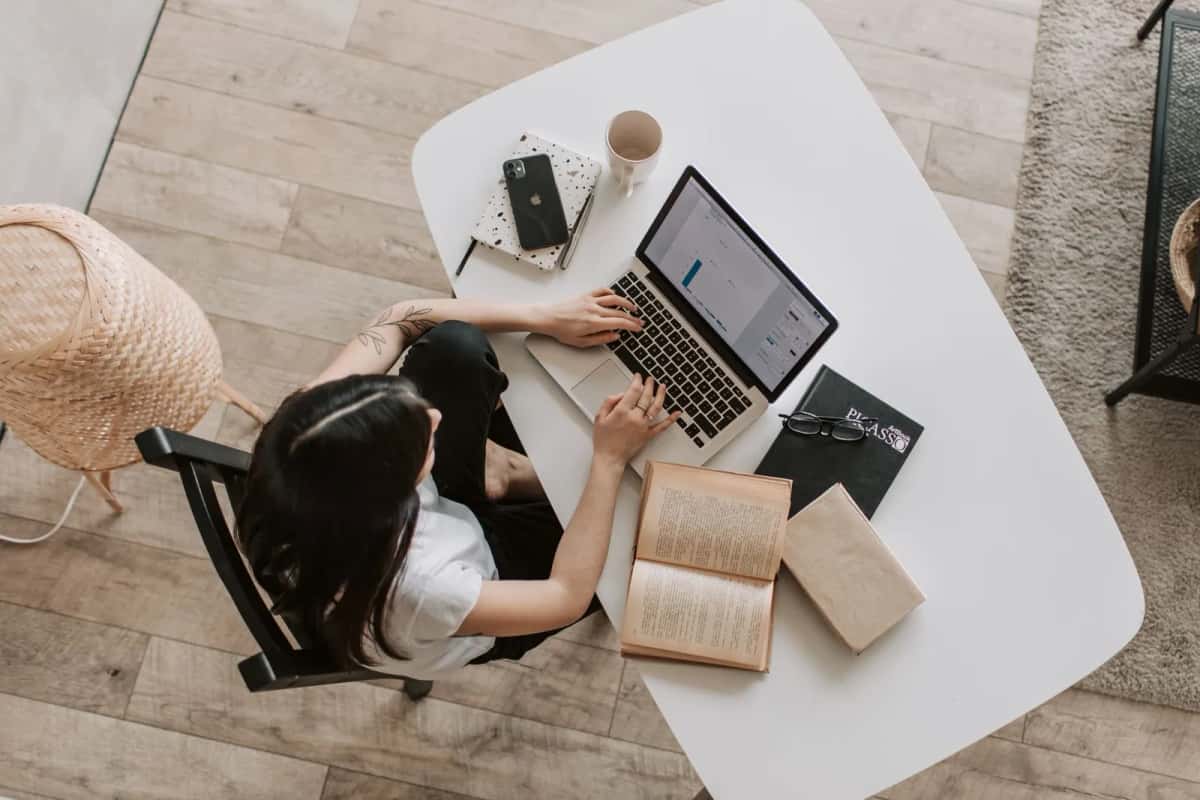 Vue de dessus d'une personne assise à une table blanche, travaillant sur un ordinateur portable à côté d'un livre ouvert et d'un carnet