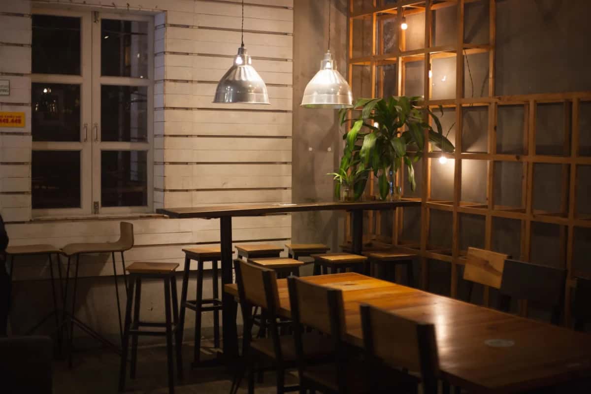 Salle de café aux murs de bois clair, tables et tabourets hauts sous deux suspensions industrielles, plante verte au fond