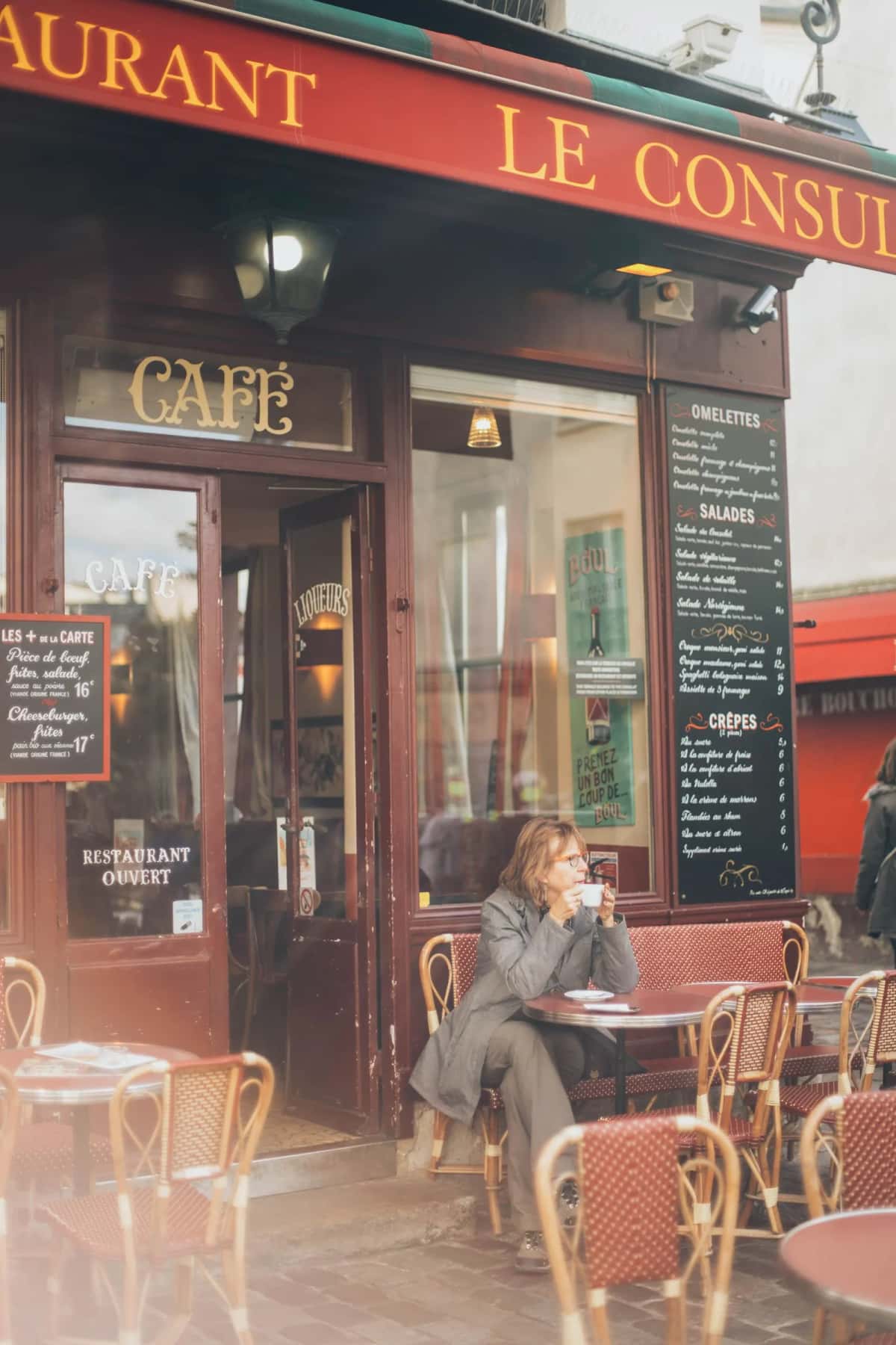 Devanture rouge d'un café-restaurant parisien avec ses tables en terrasse et ses chaises en rotin, une cliente attablée seule