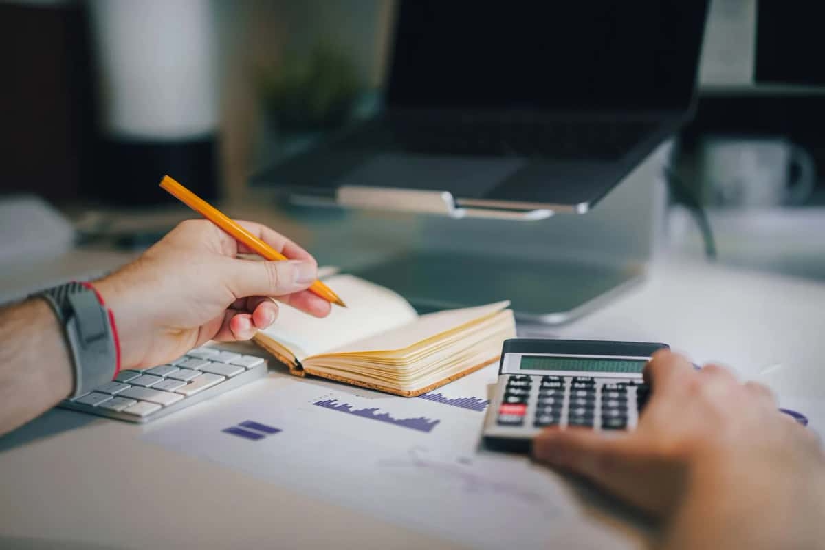 Deux mains posées sur une calculatrice et un carnet, devant un ordinateur portable et une feuille de graphiques