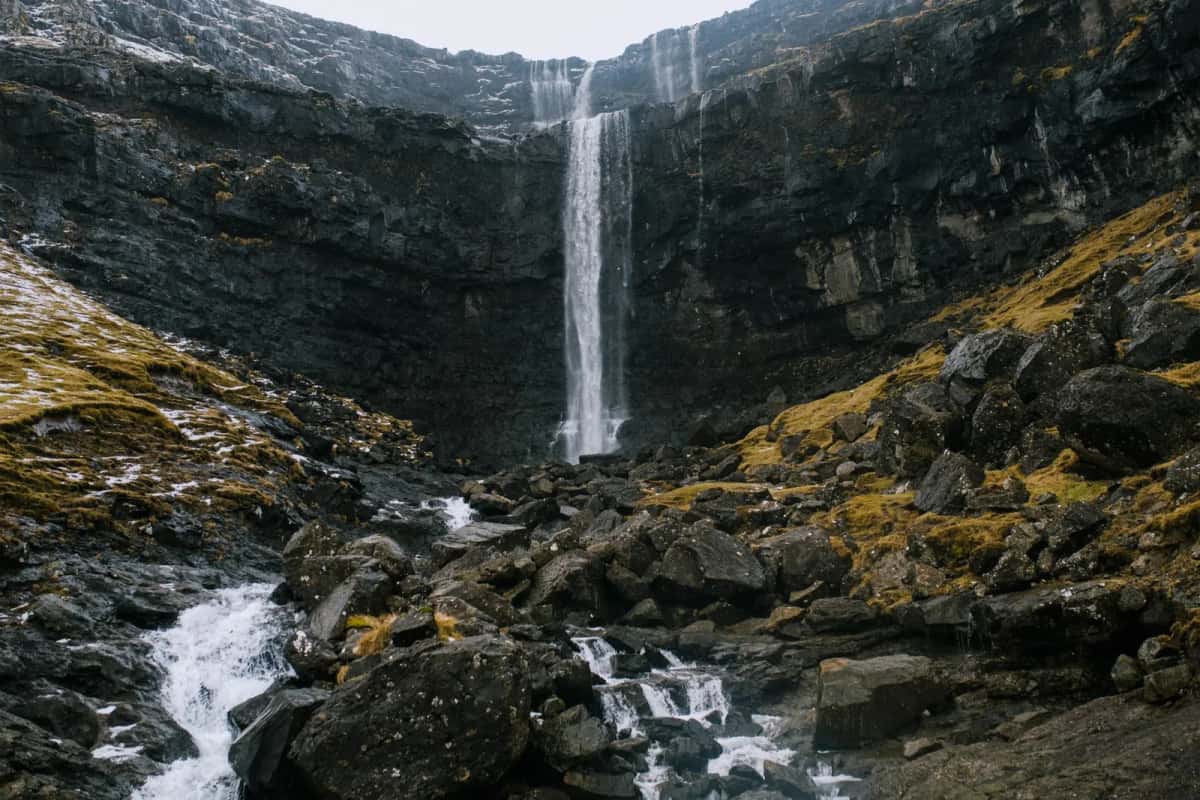 Cascade tombant en un long filet blanc dans une gorge de basalte noir tapissée de mousse et d'herbe jaune