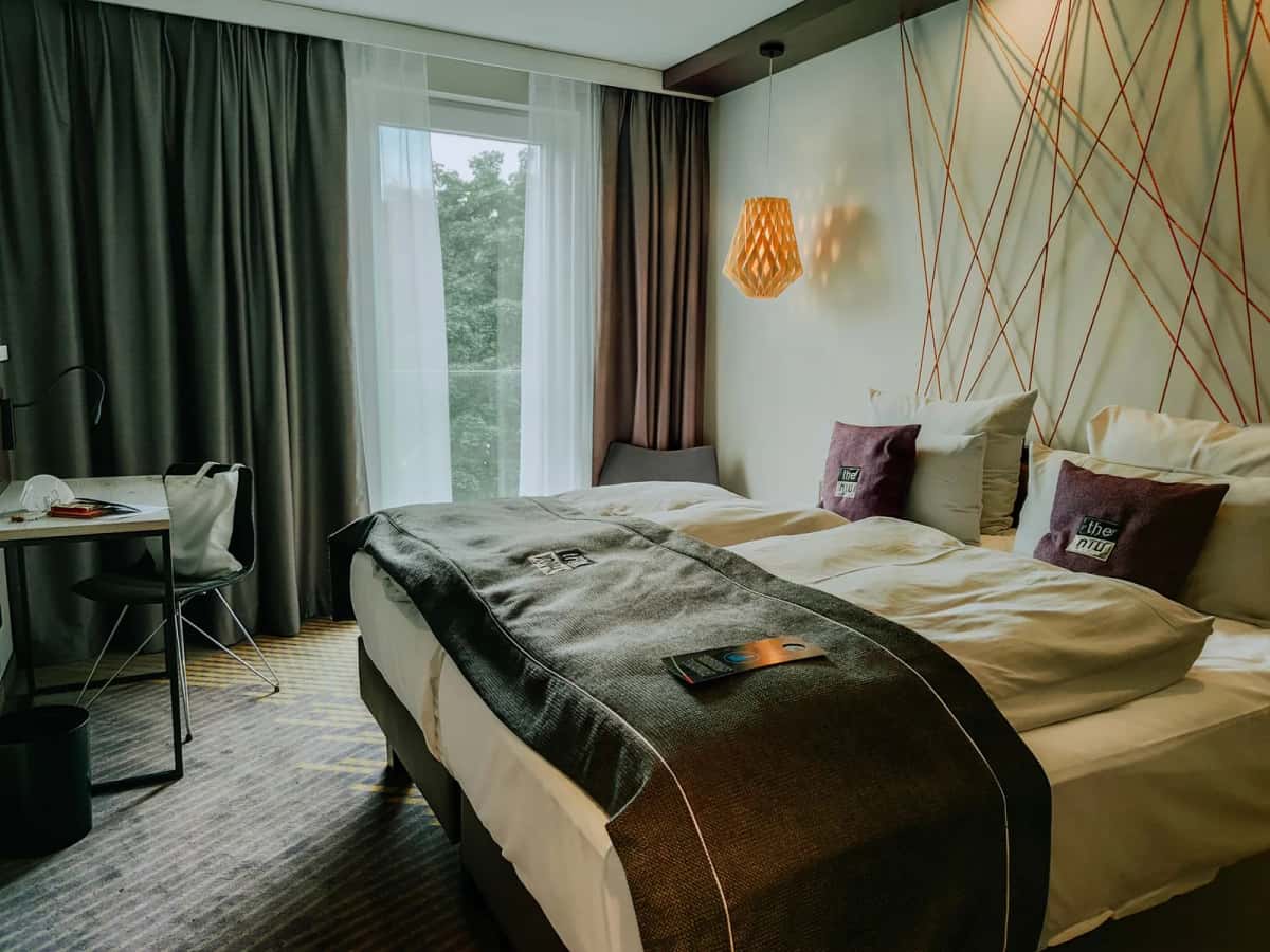 Chambre d'hôtel contemporaine, grand lit fait, bureau et chaise devant une baie vitrée aux rideaux vert foncé