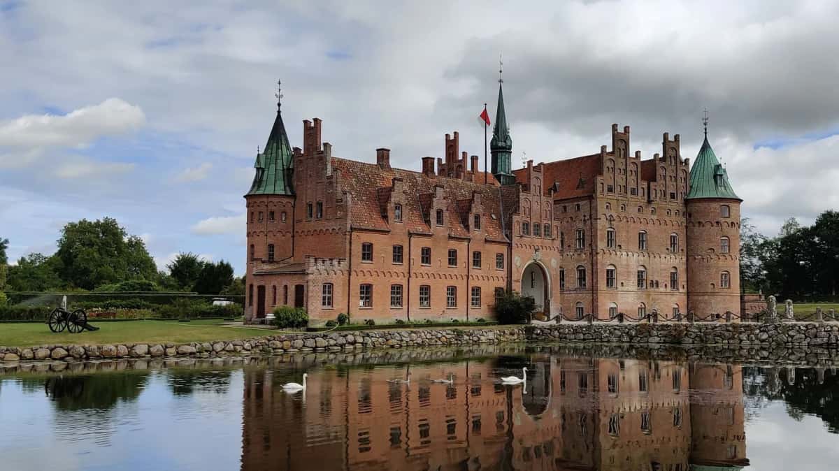 Le château d'Egeskov en Fionie, longue façade Renaissance de brique rouge à tourelles de cuivre vert, reflétée dans ses douves où nagent des cygnes