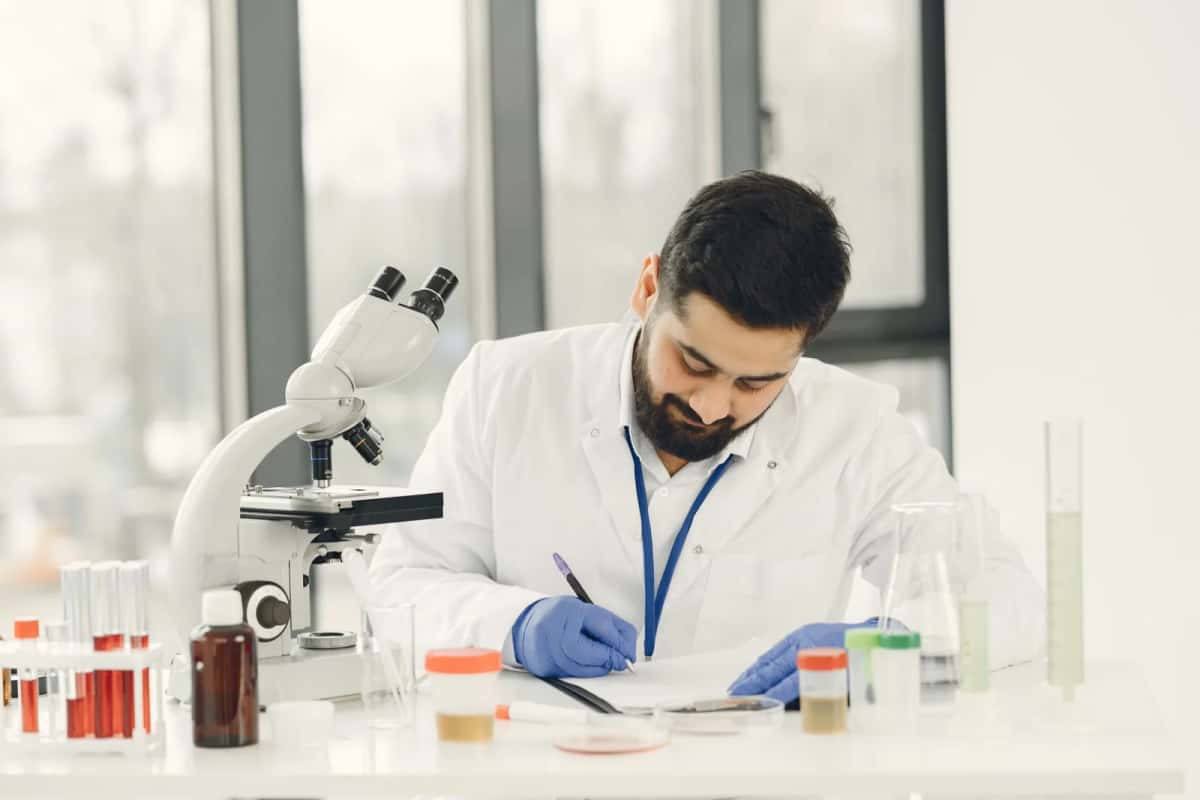 Chercheur en blouse blanche et gants penché sur un microscope, devant des portoirs de tubes à essai et d'échantillons étiquetés