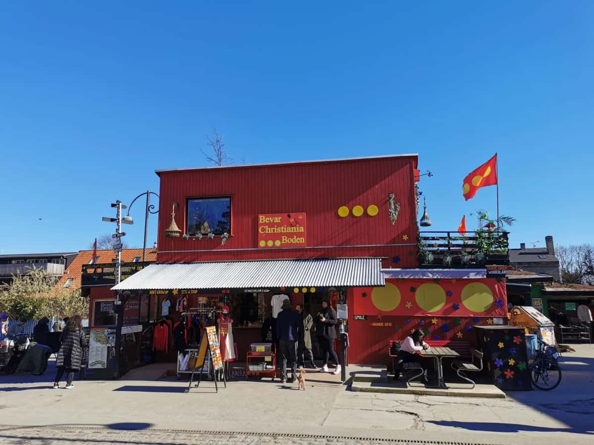 La boutique Bevar Christiania Boden, façade rouge, sous le drapeau de Christiania à trois points jaunes, avec des visiteurs attablés devant