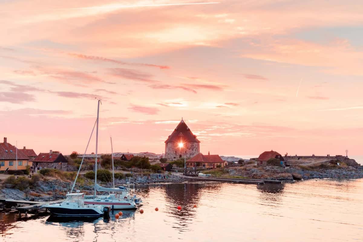 Coucher de soleil sur un chenal de port entre deux îlots de granit, tour ronde en pierre à toit conique, maisons basses à toits rouges et voiliers amarrés