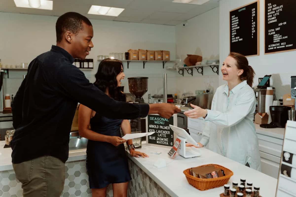 Client debout au comptoir d'un petit commerce pendant que la vendeuse encaisse derrière une caisse tactile