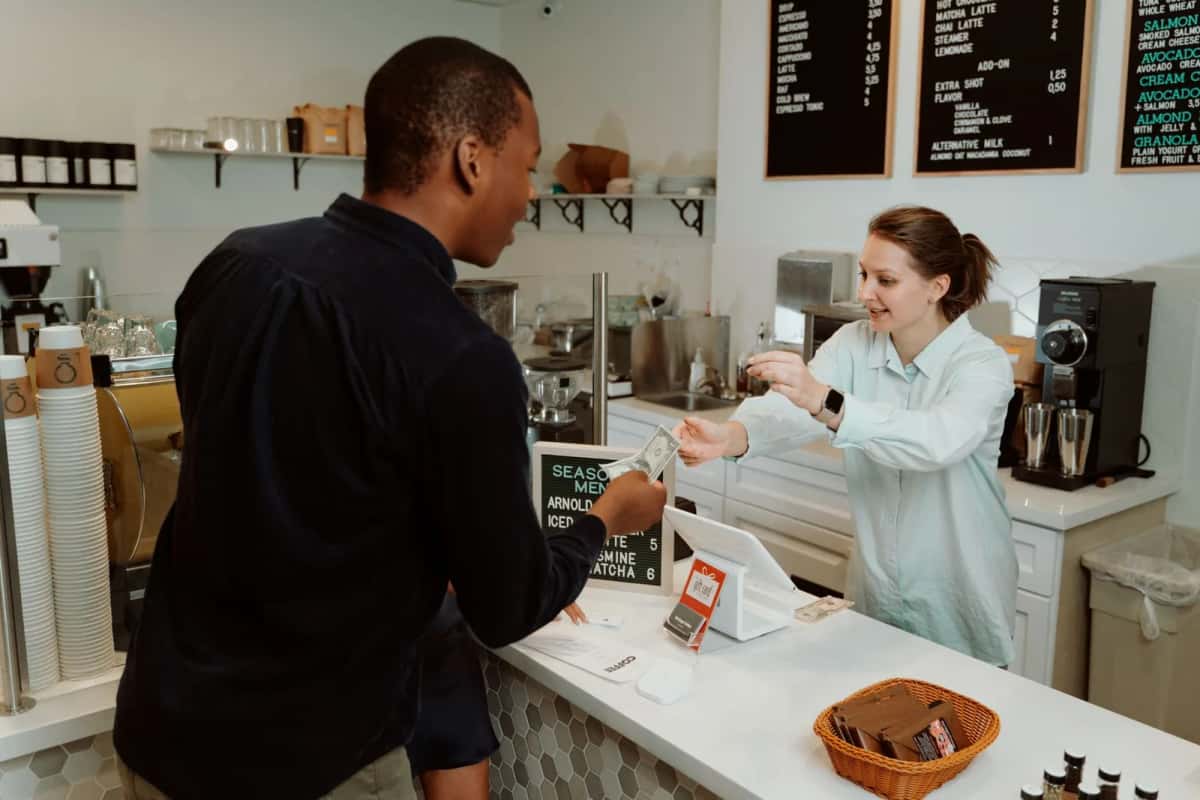 Client réglant sa commande à une serveuse derrière le comptoir d'un café, devant une ardoise de menu et une machine à espresso