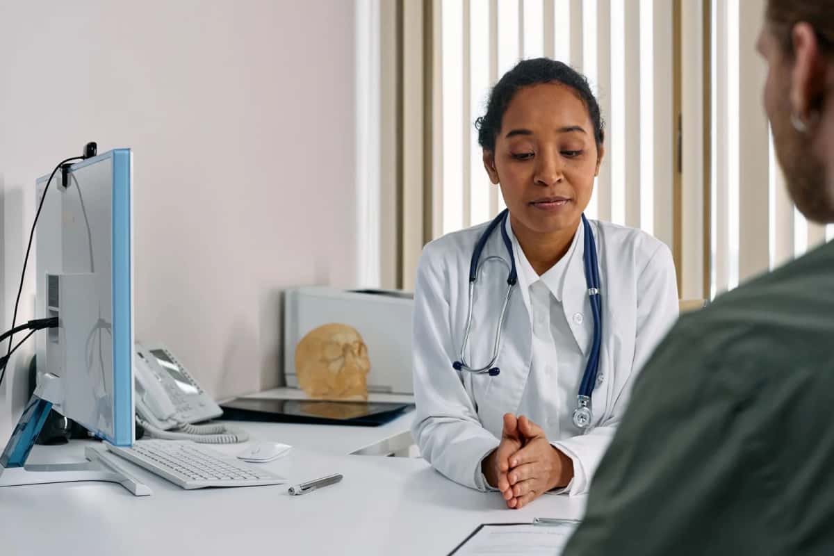 Médecin en blouse blanche assise à son bureau devant un écran, face à un patient vu de dos