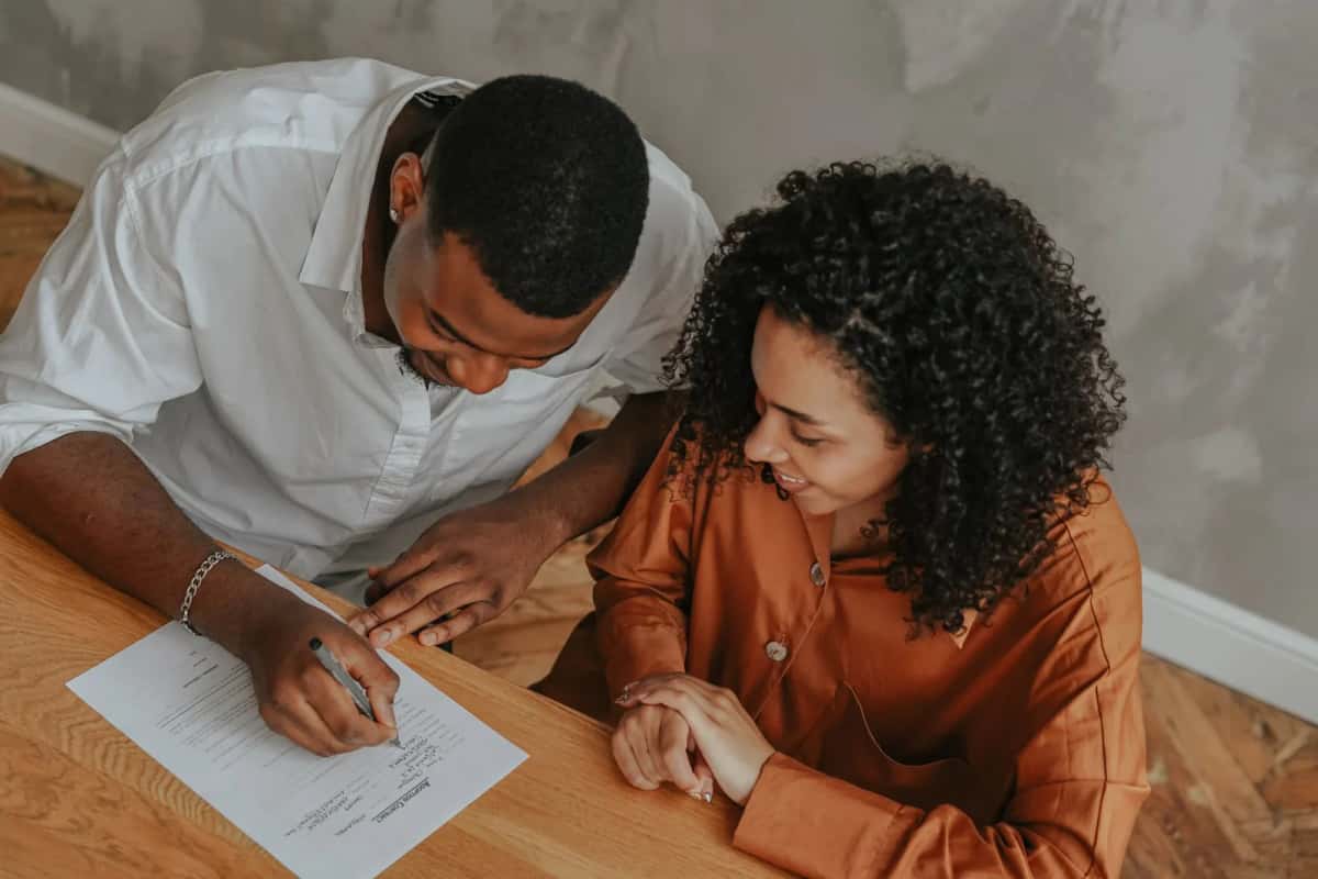 Un homme signe une feuille posée sur une table en bois pendant qu'une femme assise à côté de lui lit le document