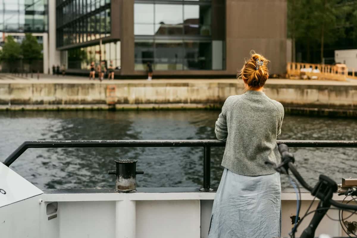 Passagère accoudée au bastingage d'un bateau-bus du port, son vélo garé à côté d'elle, façades vitrées sur la rive
