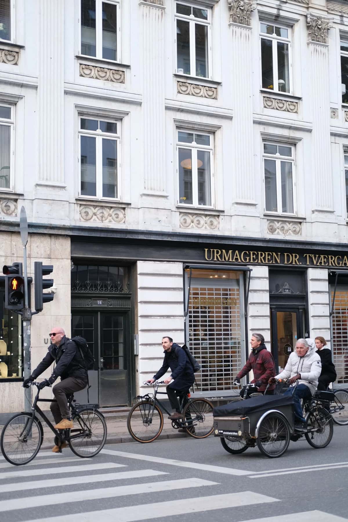 Quatre cyclistes arrêtés côte à côte à un feu, sur une piste cyclable large longeant des immeubles clairs à Copenhague