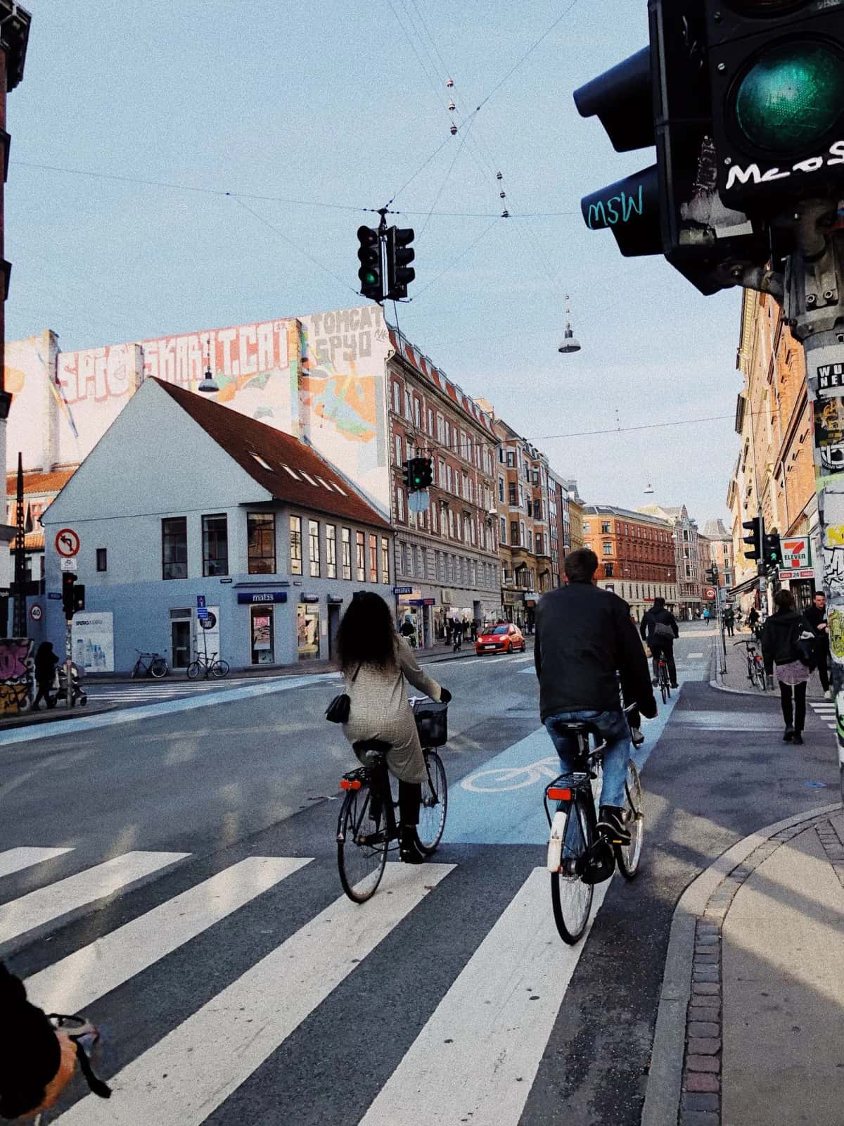 Deux cyclistes traversant un passage pour vélos peint au sol, devant un feu tricolore et un immeuble d'angle à Copenhague