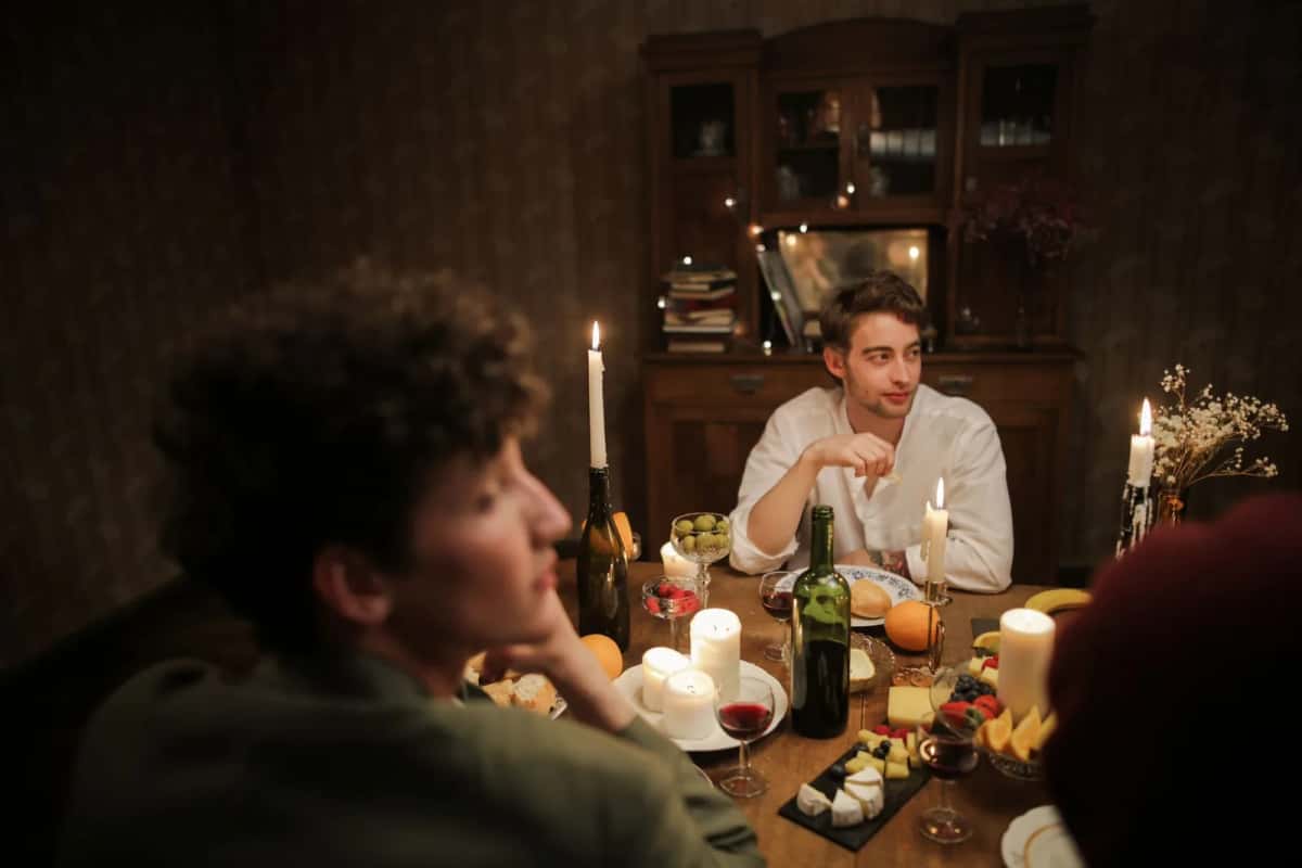 Dîner entre amis à la maison, table couverte de bougies allumées, bouteilles et petits plats, deux convives en pleine conversation