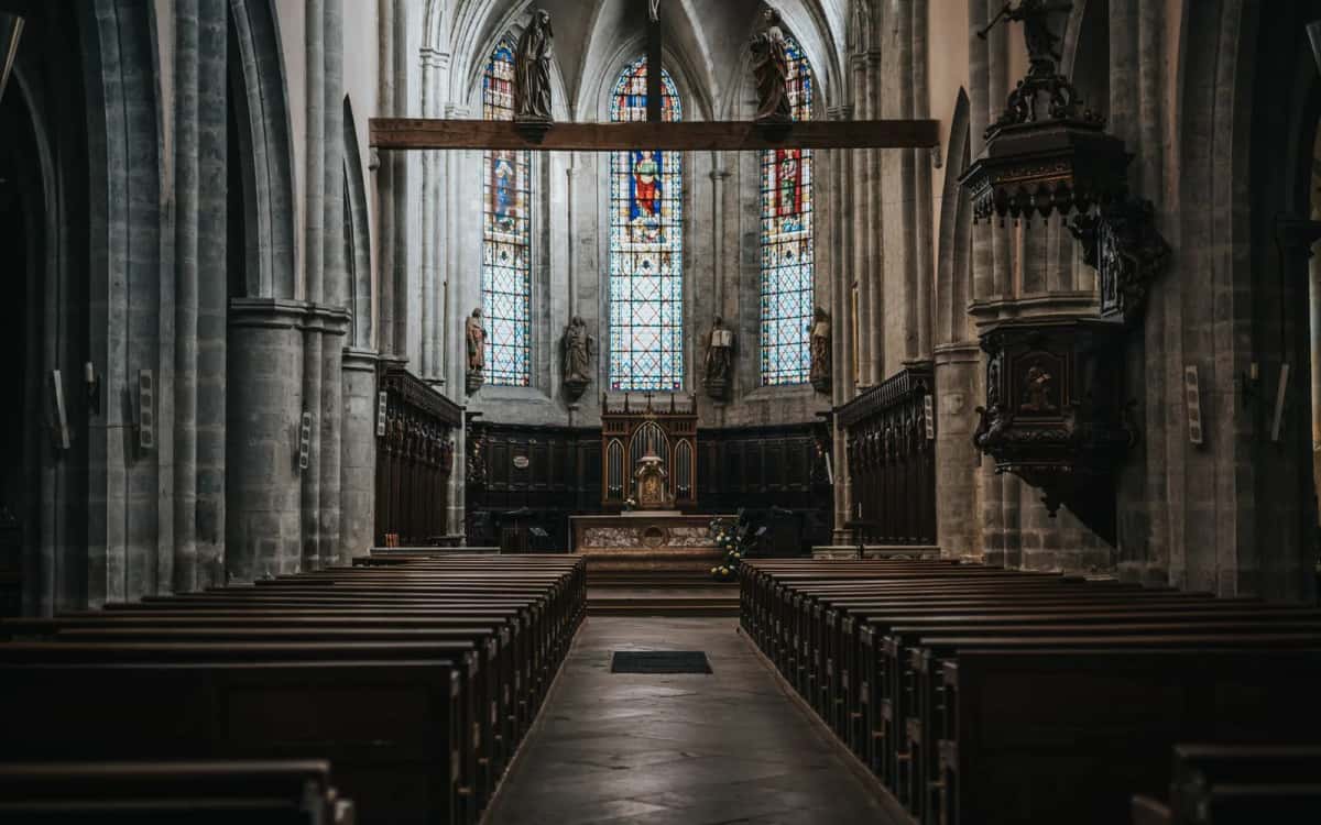 Intérieur sombre d'une église ancienne aux hautes voûtes de pierre, allée centrale bordée de bancs et vitraux au fond du chœur
