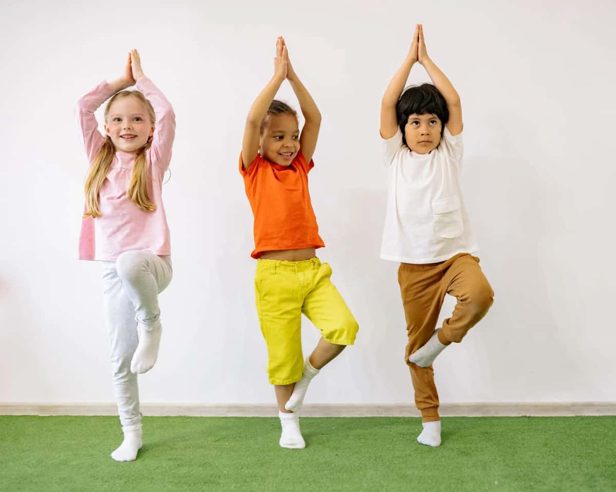 Trois jeunes enfants debout sur un tapis vert, un pied replié et les bras levés, dans une salle d'éveil aux murs blancs
