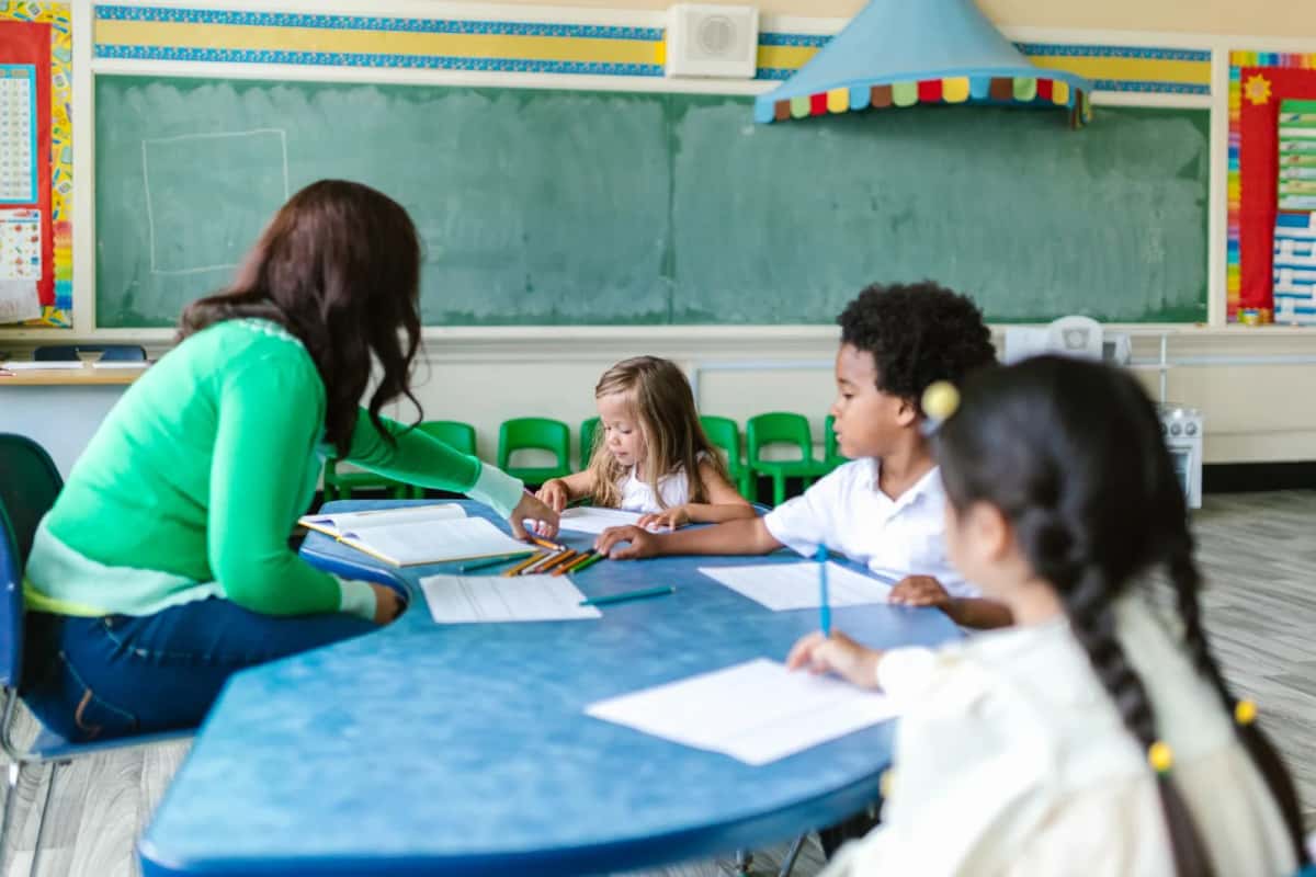 Enseignante penchée sur une table ronde autour de laquelle quatre élèves de primaire travaillent ensemble, tableau vert au fond