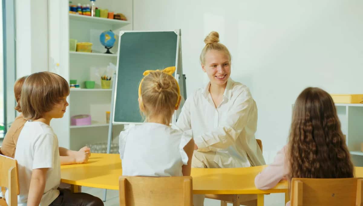 Une adulte assise à une table basse avec trois enfants de trois à cinq ans, autour d'un tableau à feuilles et d'étagères de jeux
