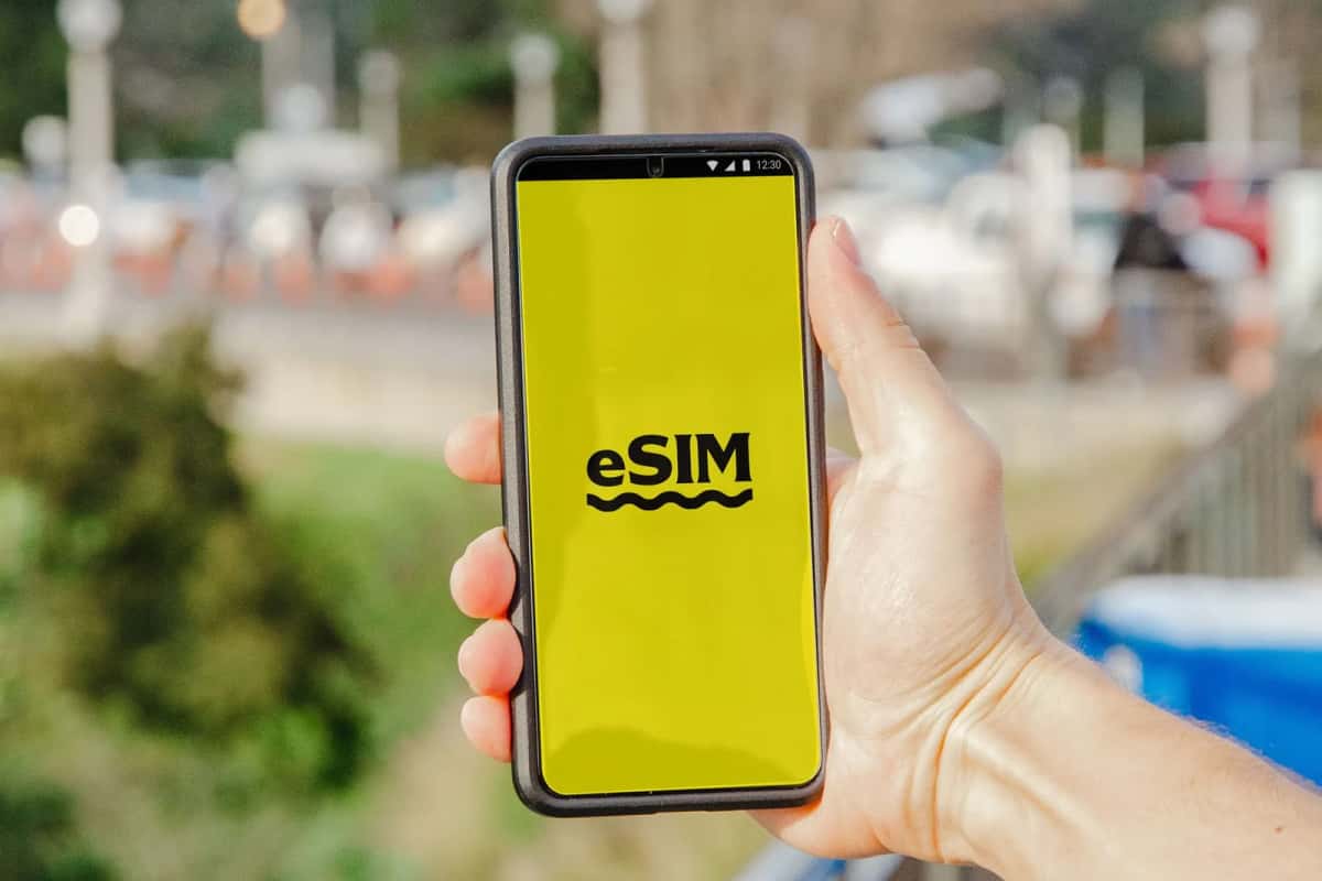 Main tenant un smartphone dont l'écran jaune affiche le mot eSIM, dans une rue en arrière-plan flou