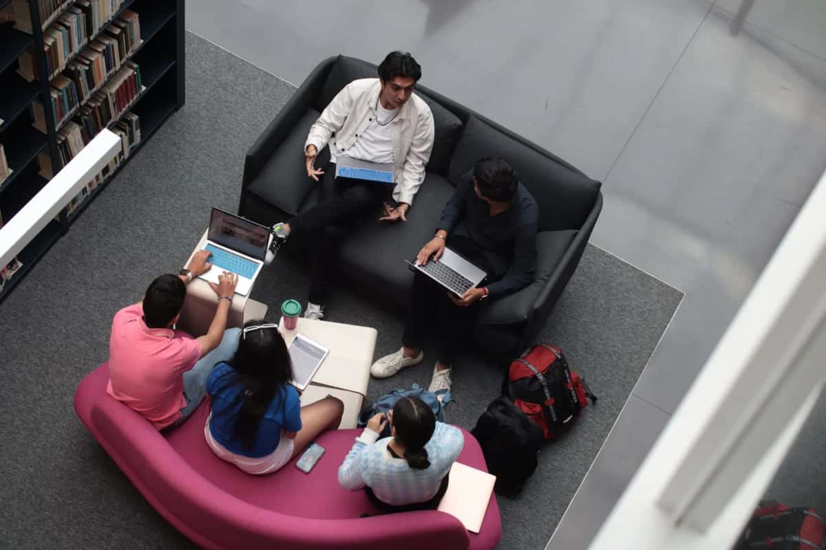 Vue plongeante sur des étudiants installés sur des canapés et un pouf rose, ordinateurs portables et cahiers ouverts, au milieu de rayonnages