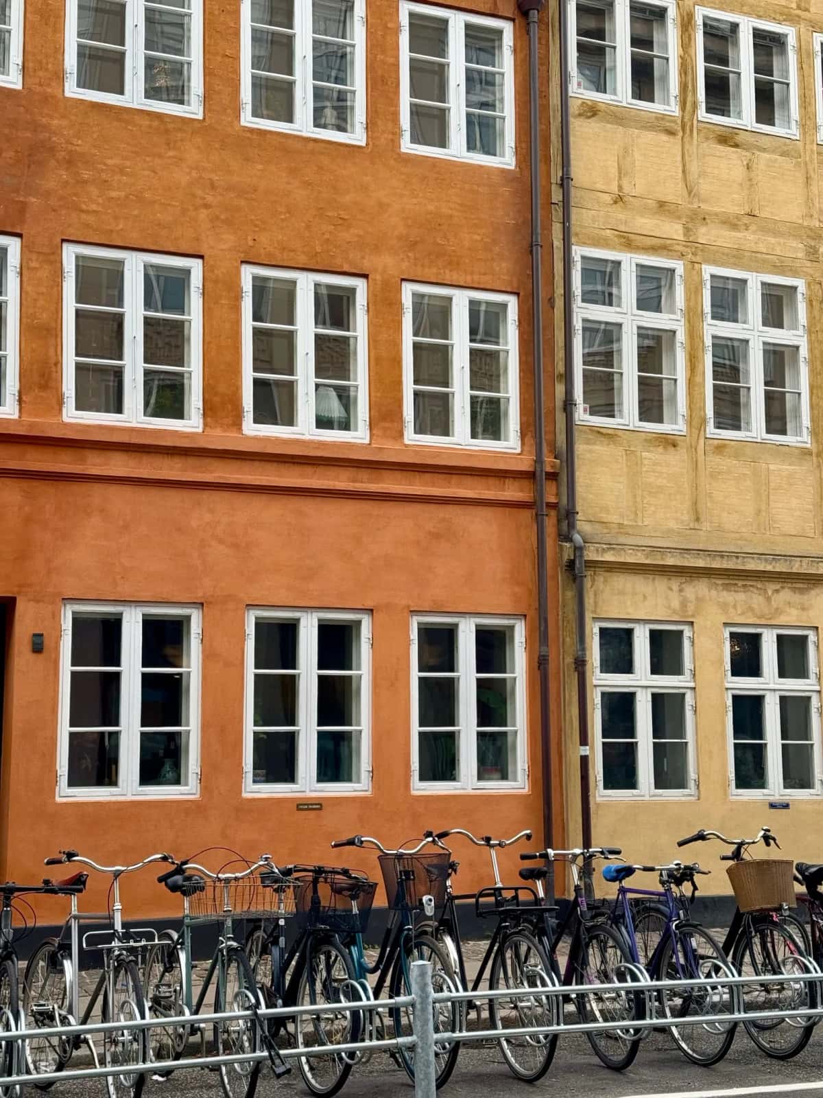 Une longue rangée de vélos garés le long d'un trottoir, devant une façade d'immeuble ocre à fenêtres blanches
