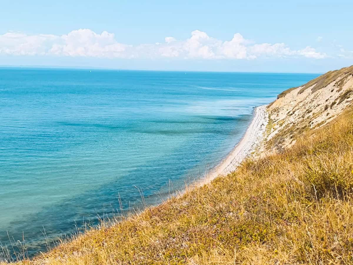 Bord de falaise claire couvert d'herbes sèches, plongeant vers une mer turquoise et un liseré de galets, sans barrière ni sentier balisé