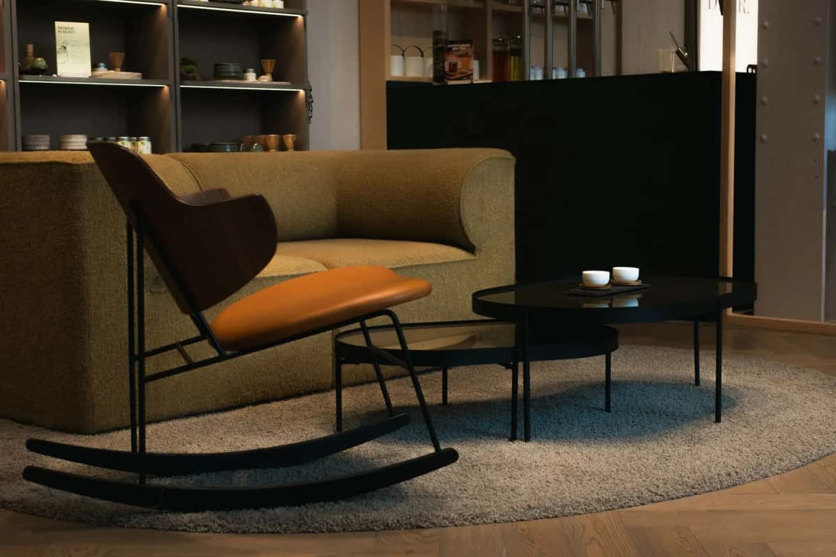Fauteuil à bascule en bois cintré et assise ocre, posé sur un tapis rond près d'une banquette basse et de tables gigognes noires