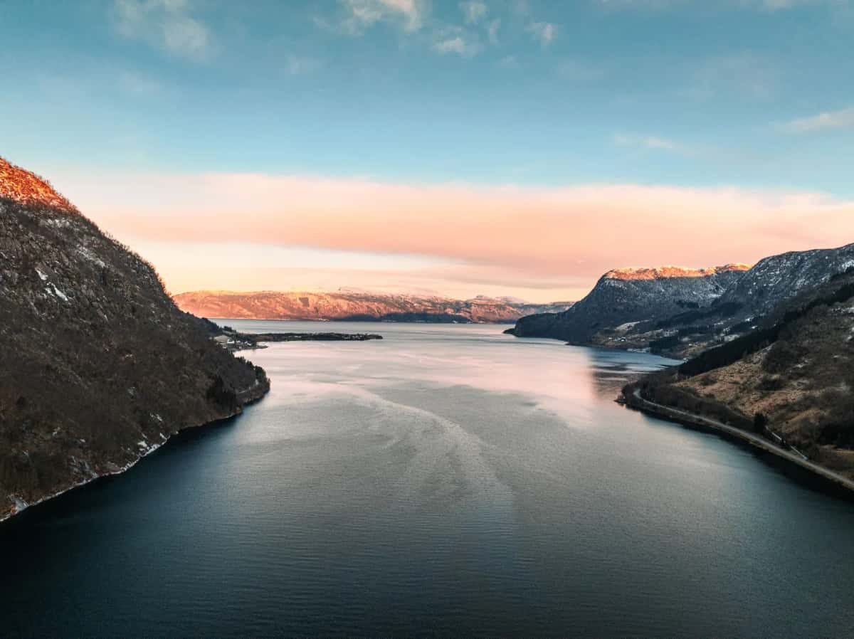 Fjord large et immobile encadré par des versants raides encore couverts de neige, dans une lumière rose de fin de journée