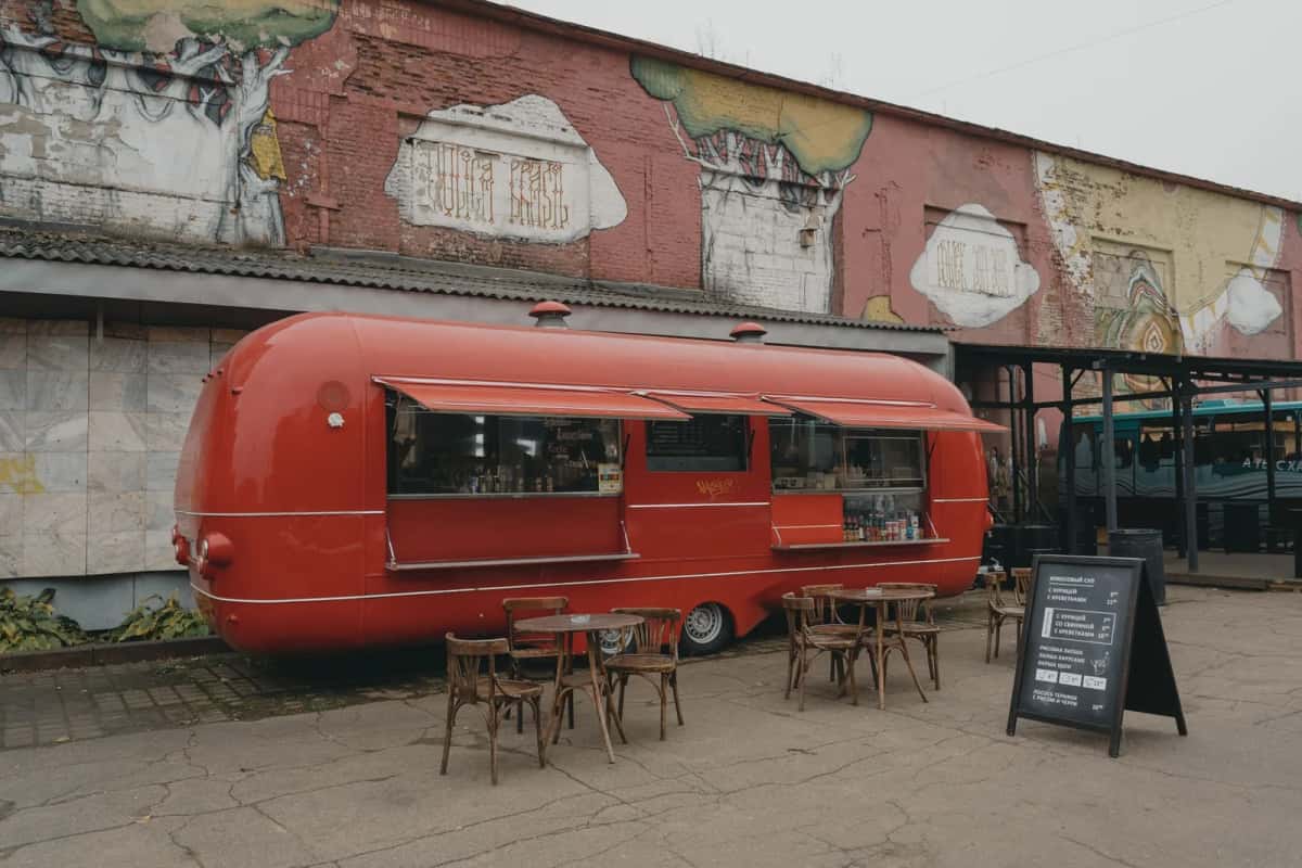 Food truck rouge à carrosserie arrondie garé devant un ancien hangar couvert de fresques, tables et tabourets en bois installés dehors sur le béton