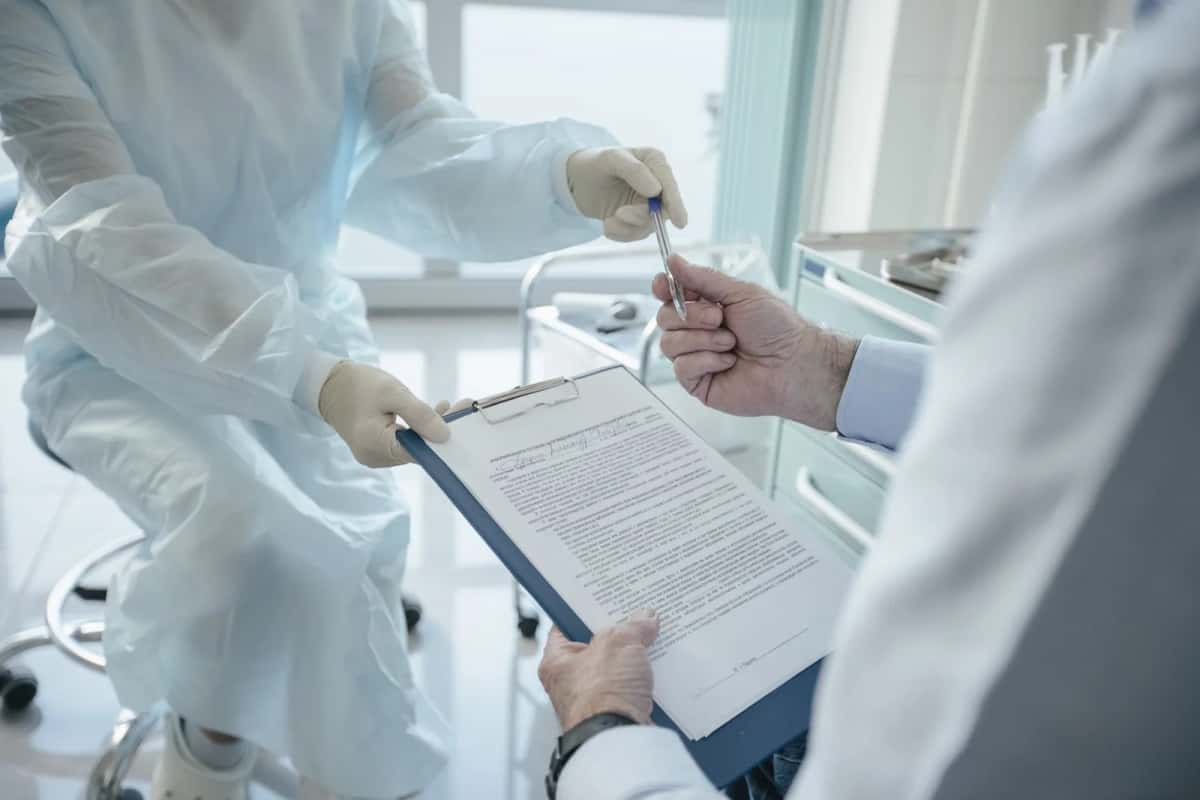 Un soignant en blouse tend un porte-documents et un stylo à un confrère en blouse blanche, dans une salle de soins