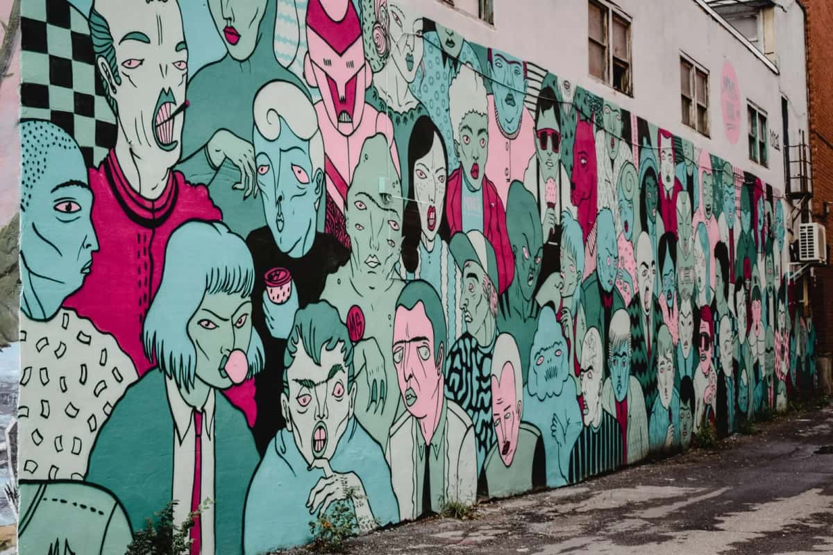 Longue fresque murale peinte sur le mur aveugle d'un bâtiment bas : des dizaines de visages stylisés en bleu, vert et rose