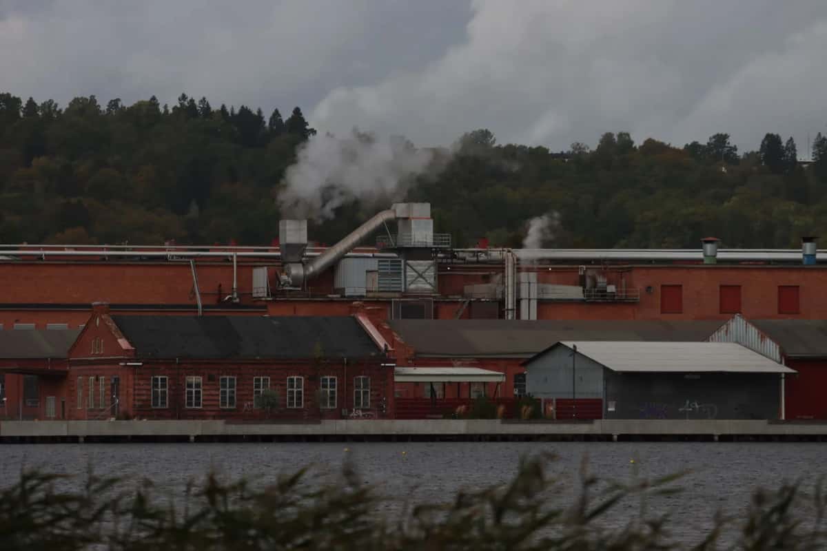 Longues halles industrielles en brique rouge alignées au bord de l'eau, panache de vapeur au-dessus des toits et colline boisée en arrière-plan
