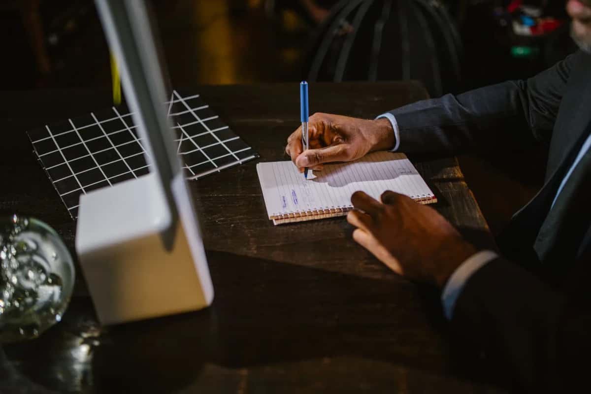 Homme en costume assis à une table sombre, un stylo à la main, prenant des notes sur un carnet à spirale sous une lampe de bureau