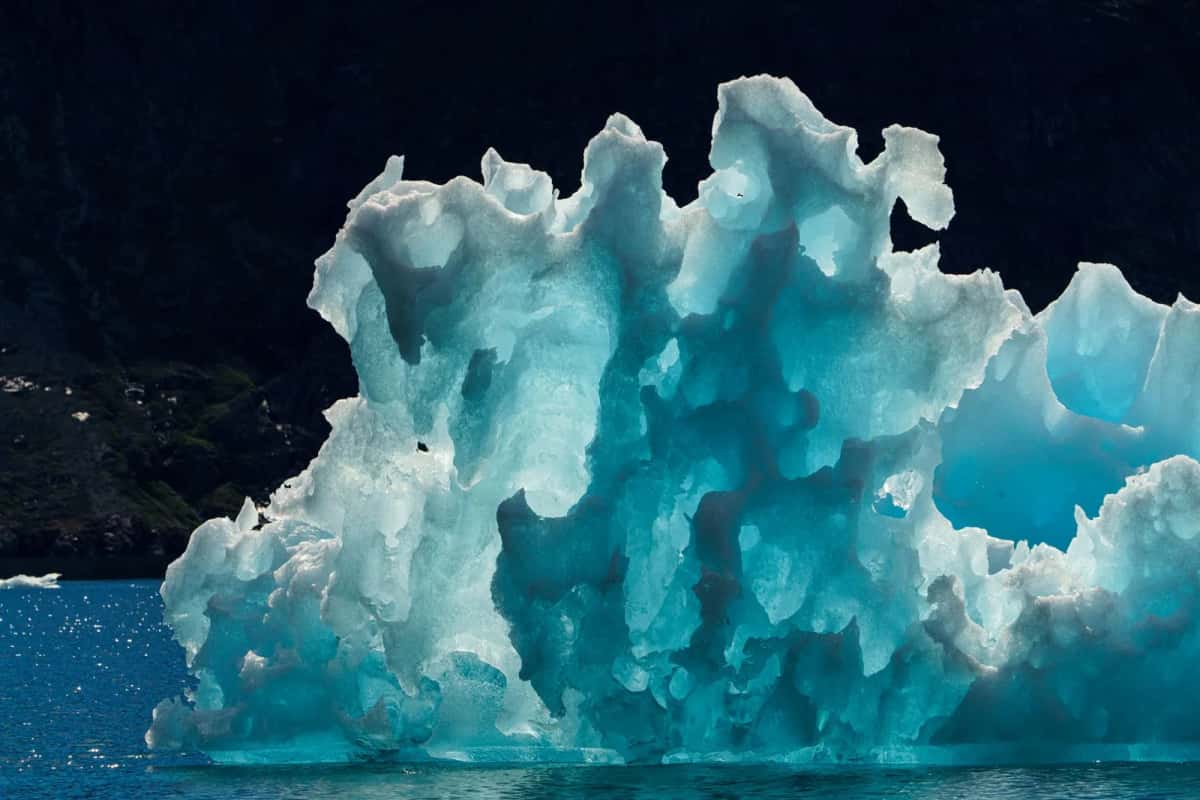 Iceberg bleu turquoise aux parois sculptées par la fonte, flottant devant une paroi rocheuse sombre