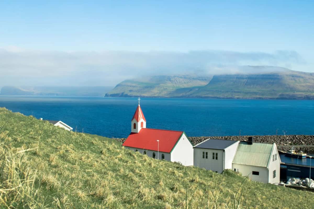 Petite église blanche à toit rouge et maisons de bois au bord d'un bras de mer, face à des montagnes au sommet plat
