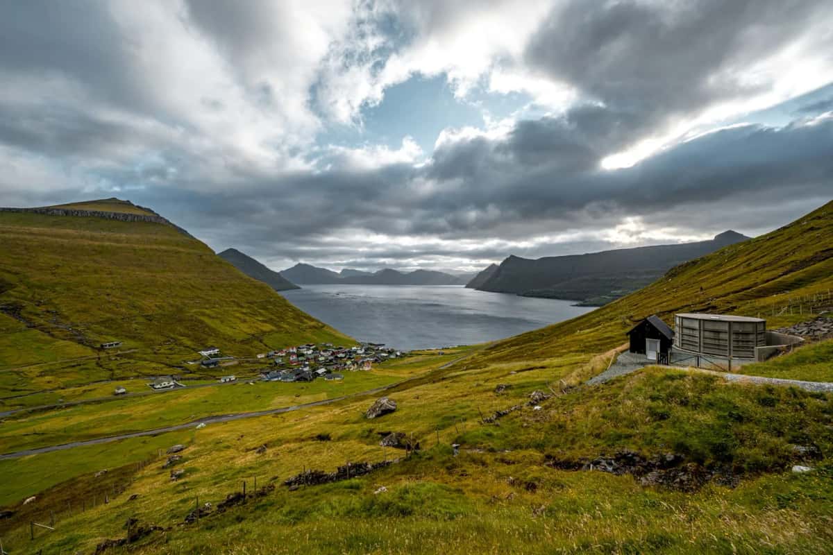 Village dispersé au fond d'un bras de mer encaissé entre des montagnes vertes, cabane à toit d'herbe au premier plan, ciel de nuages bas