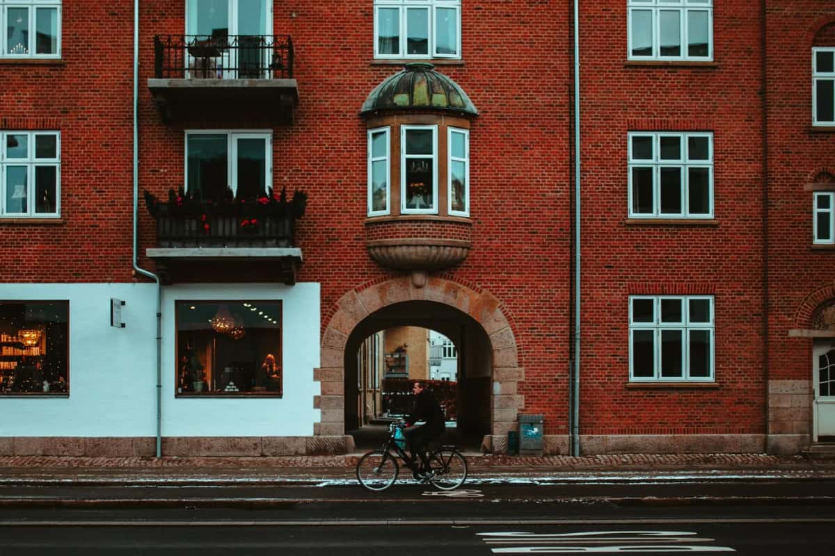 Cycliste longeant une façade de brique rouge à Aalborg, porche voûté au centre, bow-window coiffé d'un dôme de cuivre et piste cyclable au premier plan
