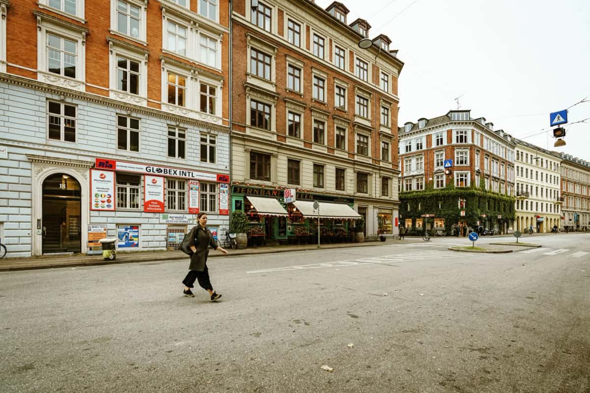 Immeubles de cinq étages aux façades rouge, ocre et vert d'eau alignés le long d'une large rue, une passante traverse la chaussée