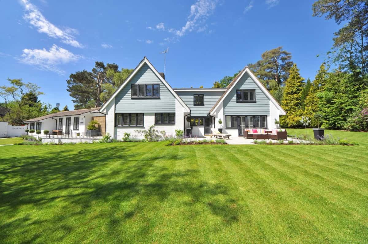 Maison scandinave à bardage blanc, à deux pignons, devant une grande pelouse tondue bordée d'arbres