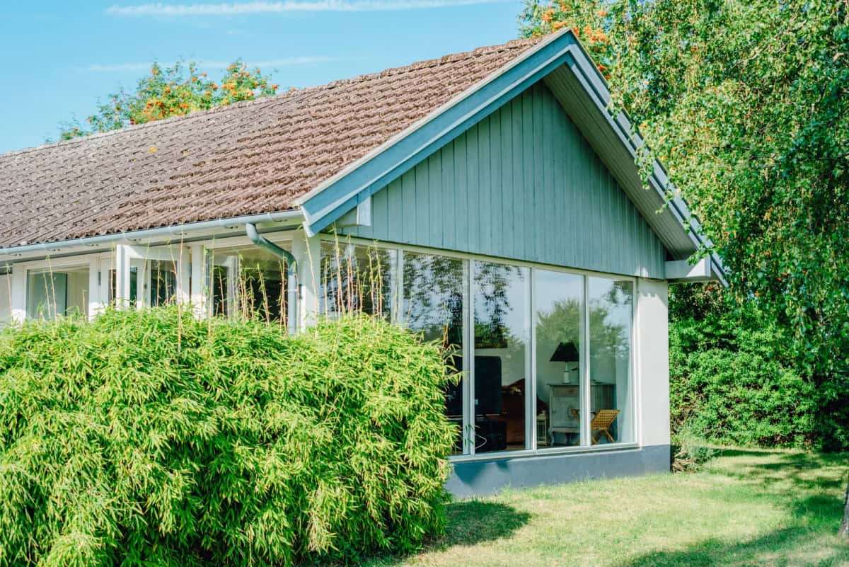 Maison individuelle en bois peint bleu-vert, avec véranda vitrée, entourée d'un jardin arboré