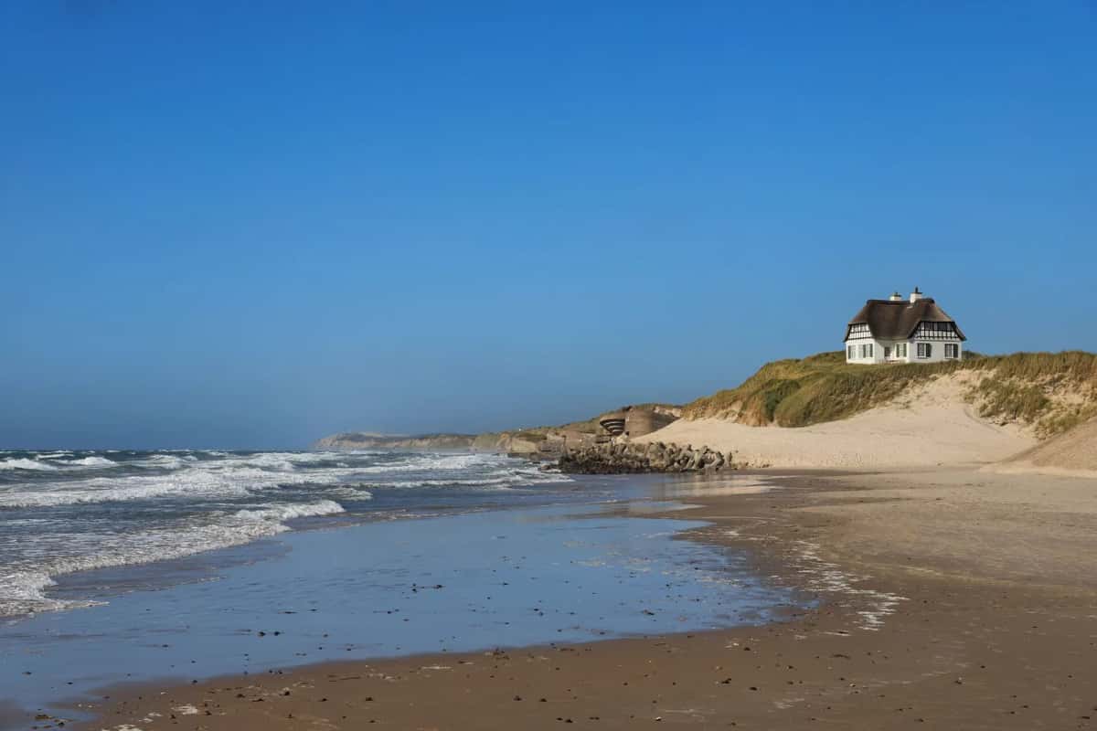 Maison blanche à toit de chaume posée au sommet d'une dune, au-dessus d'une longue plage battue par les vagues