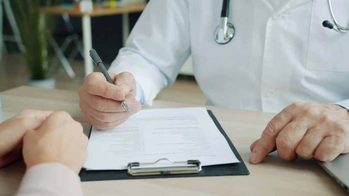 Un médecin en blouse blanche remplit un formulaire sur un porte-bloc, les mains du patient posées sur la table en face