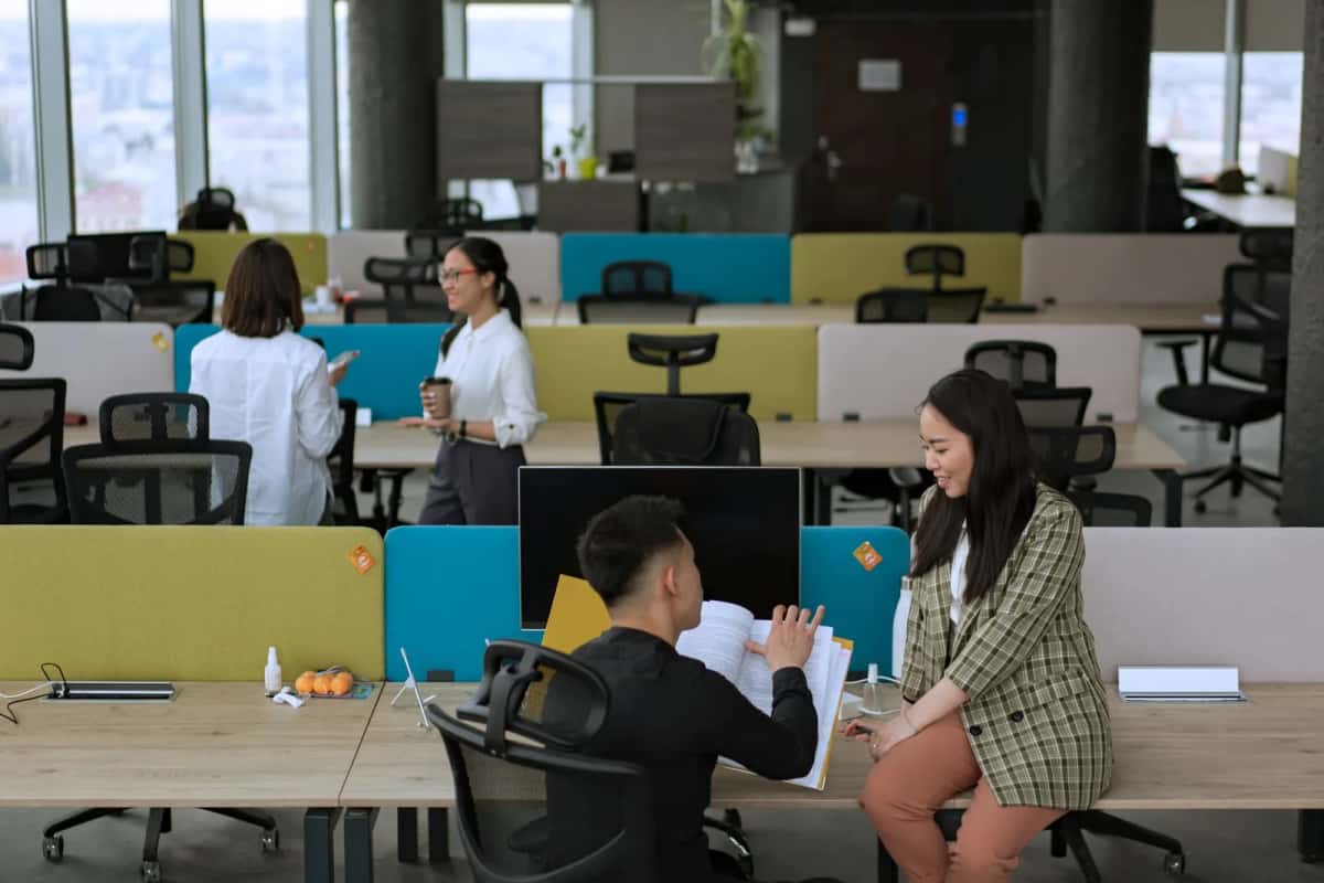 Open space aux cloisons basses colorées, deux collègues discutent debout près d'un poste de travail pendant que d'autres travaillent assis