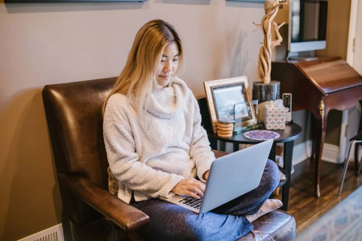 Femme installée dans un fauteuil de salon, ordinateur portable ouvert sur les genoux, près d'un secrétaire en bois