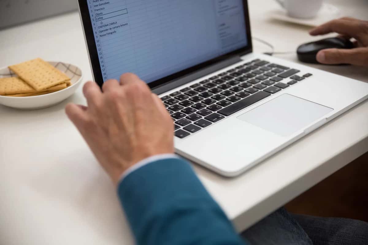 Mains posées sur un ordinateur portable ouvert sur un tableur, sur un bureau blanc, à côté d'une tasse et d'une soucoupe de biscuits